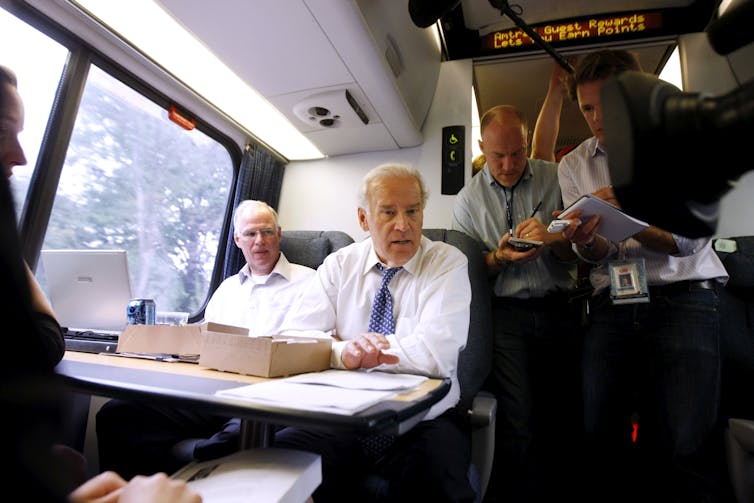 Biden, seated with aides, speaks to reporters standing in the train aisle.