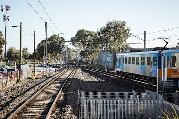 Railway tracks extend into the distance with a blue-and-white passenger train on the right; overhead lines, fencing, trees, and a clear sky.