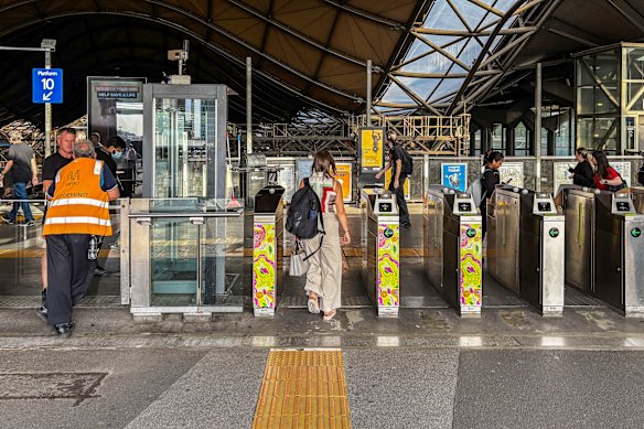 Passengers pass through ticket gates at a modern train station; a worker in an orange vest stands to the left near a glass elevator.