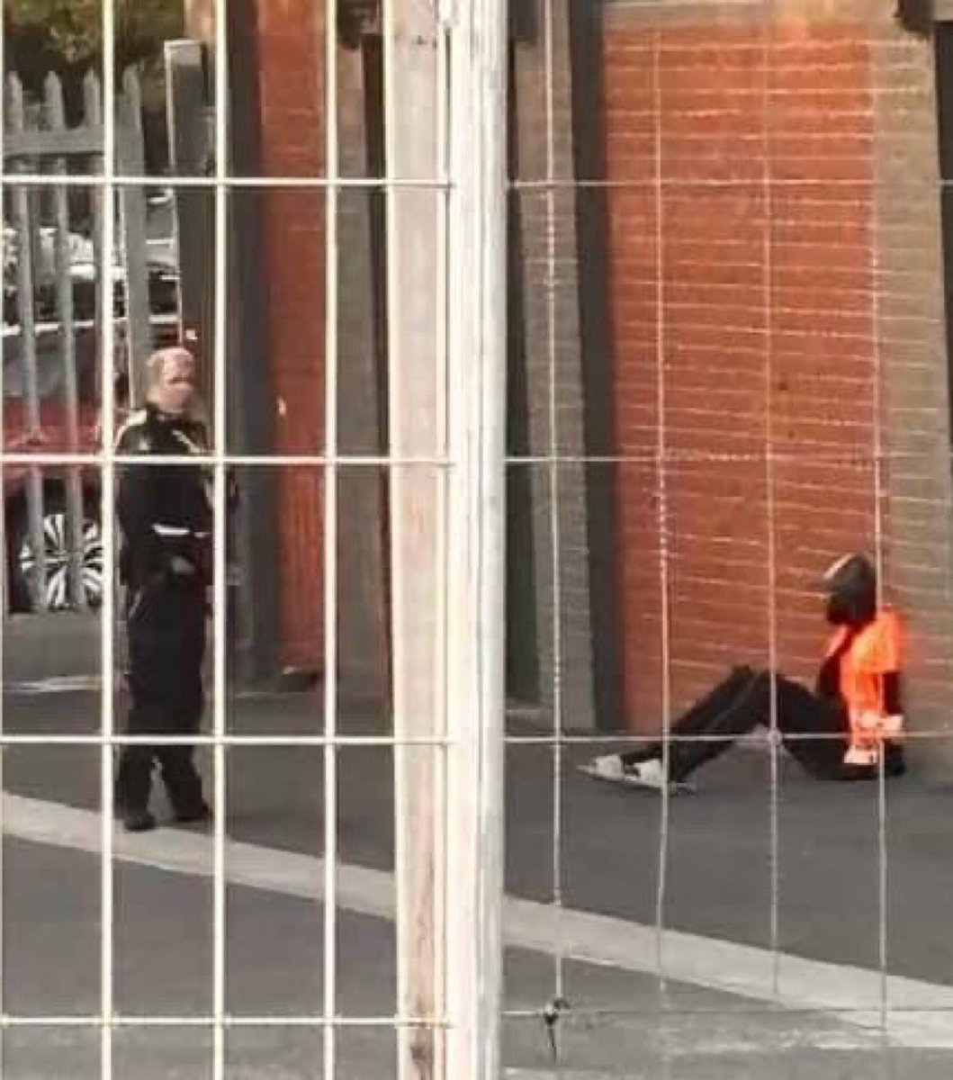 Person in an orange safety vest sits on the sidewalk near a brick wall, with another person in dark clothing standing nearby behind a glass facade.