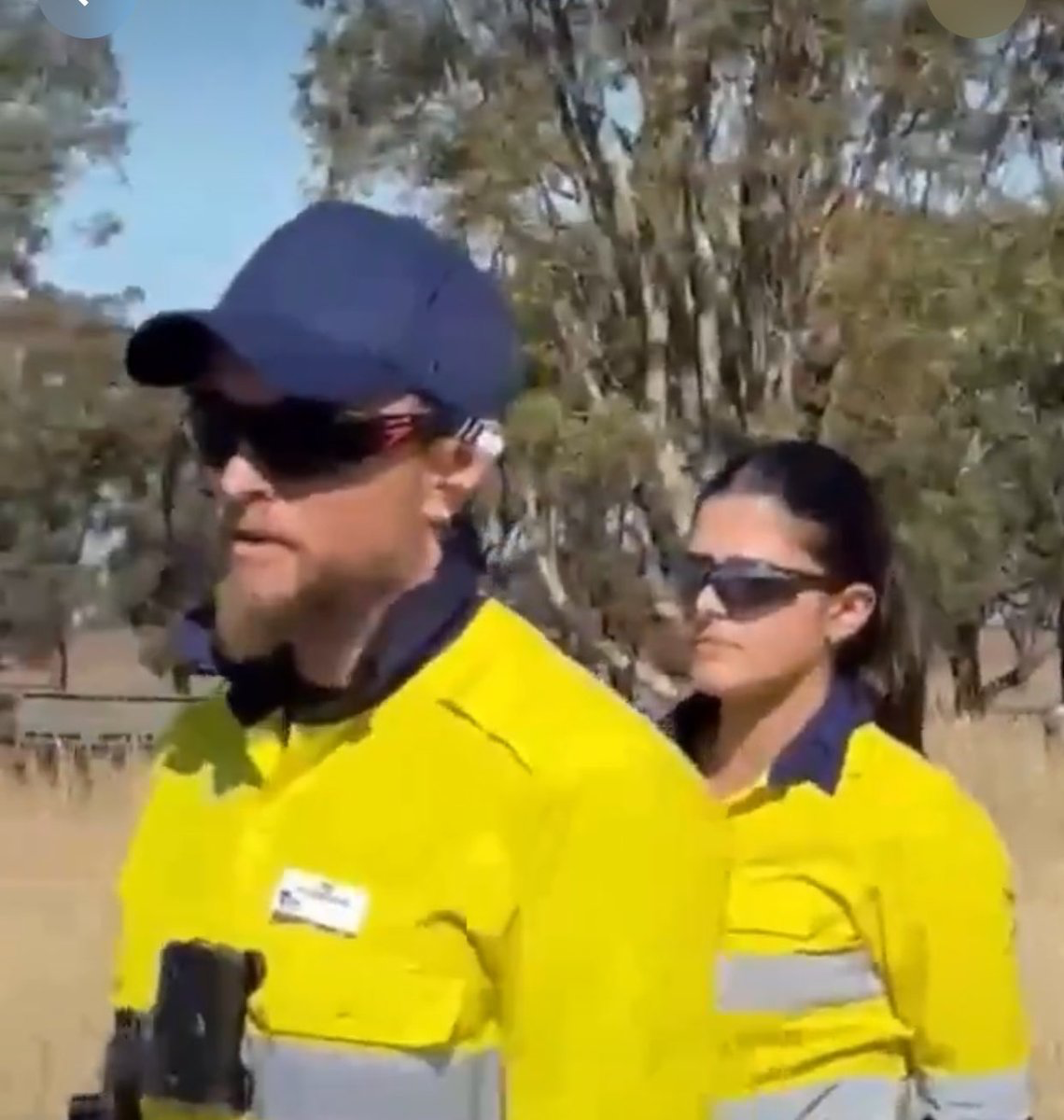 Two workers in bright yellow safety jackets wearing sunglasses walk outdoors in a sunny, rural area.