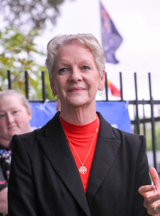 Older woman with short gray hair in a red top and black blazer, standing outdoors near a fence with flags, smiling at the camera.