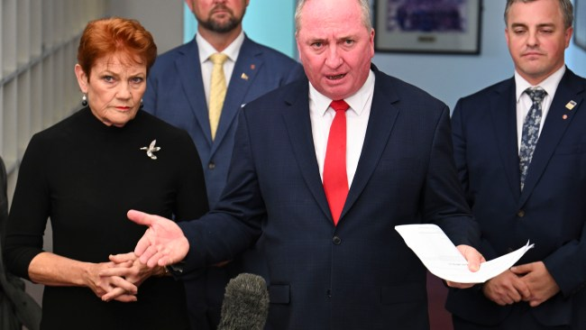 Man in a dark suit and red tie speaks at a press conference, holding papers, with a woman in black beside him and two suited men behind.