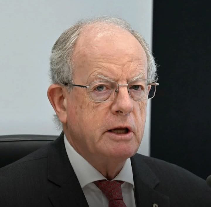 Older man with gray hair and glasses, wearing a dark suit and red tie, speaking or addressing an audience in a formal setting