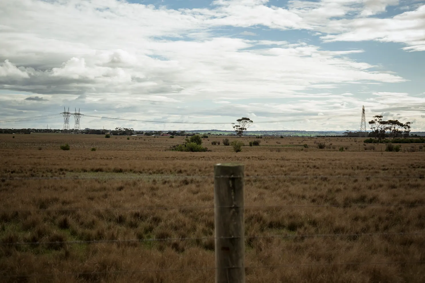 Open, dry grass field with a wooden fence post in the foreground and power lines across the horizon.