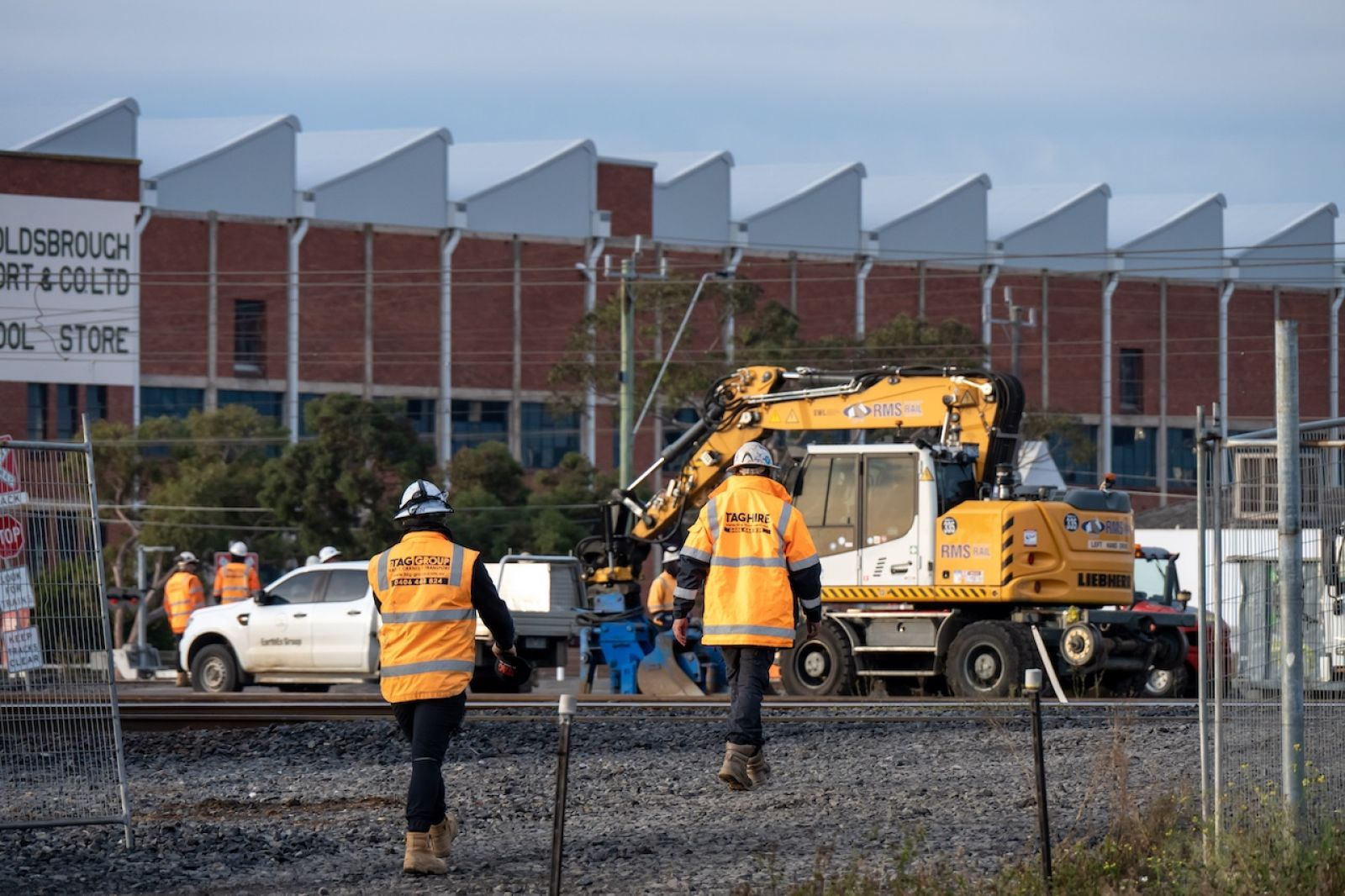Construction site beside railroad tracks with workers in orange safety vests and a yellow excavator in the background
