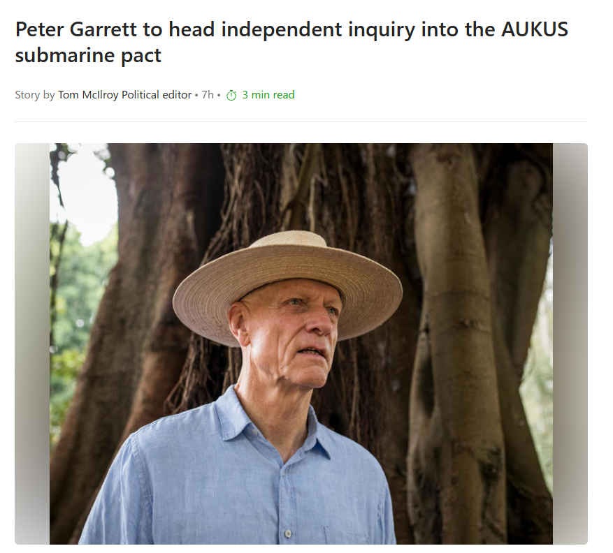 Older man wearing a straw hat and light blue shirt stands outdoors in front of thick tree roots.