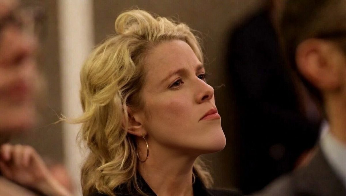 Blonde woman in profile with hoop earring, looking upward in a dim indoor setting as blurred figures loom in the background.