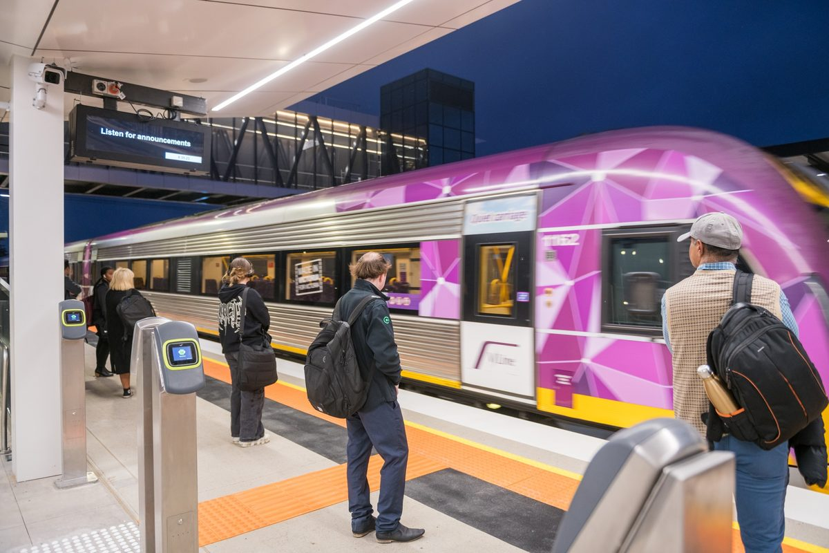 Passengers stand on a platform beside a purple geometric-patterned train as it stops to board.