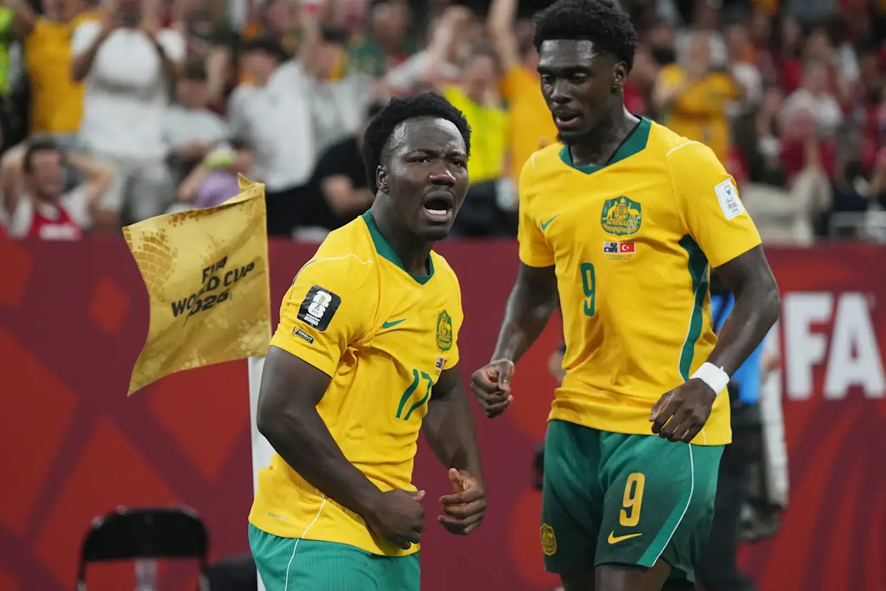 Two soccer players in yellow and green celebrate on the field as fans cheer in the stands behind them.