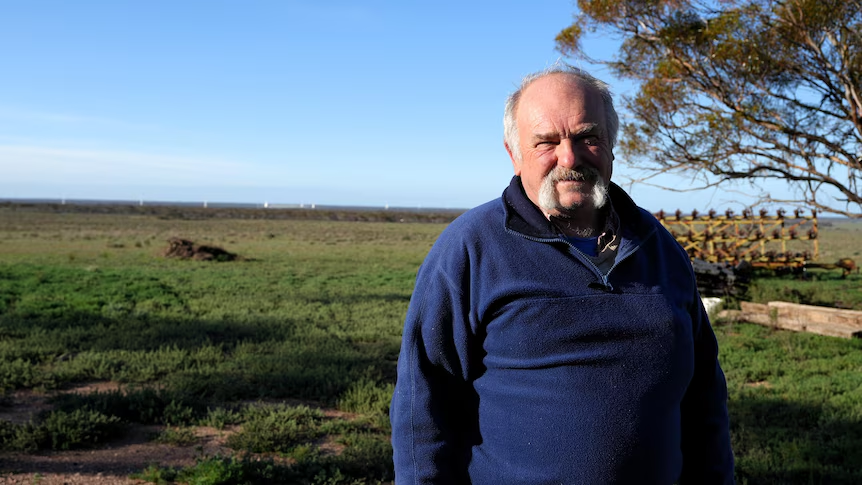 Older man with a white moustache wearing a blue fleece stands in a grassy rural field under a clear blue sky.