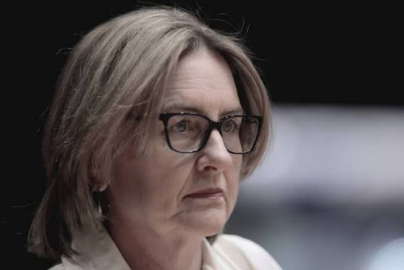 Close-up of a mature woman with short blonde hair and black-framed glasses, looking to the left with a neutral expression.