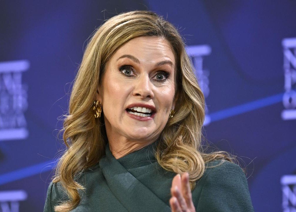 Woman with blonde hair speaking at a panel, gesturing with her right hand, wearing a dark green outfit and gold earrings in front of a blue backdrop.
