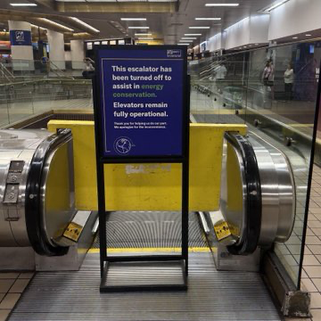 Blue sign on a closed escalator at a transit station, noting the escalator is off to save energy and that elevators remain fully operational.