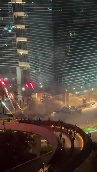 Nighttime city scene with a glass high-rise, a curved elevated walkway, and visible sparks and smoke near the building.