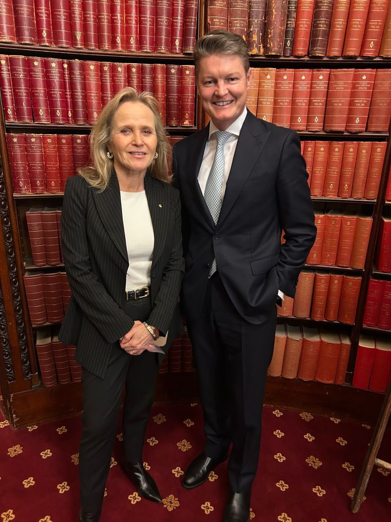 Two professionally dressed people posing for a photo in front of tall red leather-bound books on library shelves in a formal setting.
