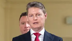Man in a dark suit and red tie speaks to the media, with another suited man visible in the background.