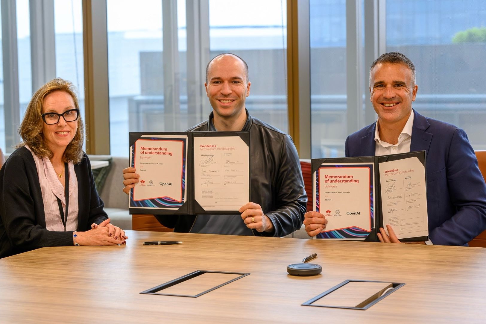 Three professionals at a conference table display signed memorandums of understanding to the camera in a bright office setting.