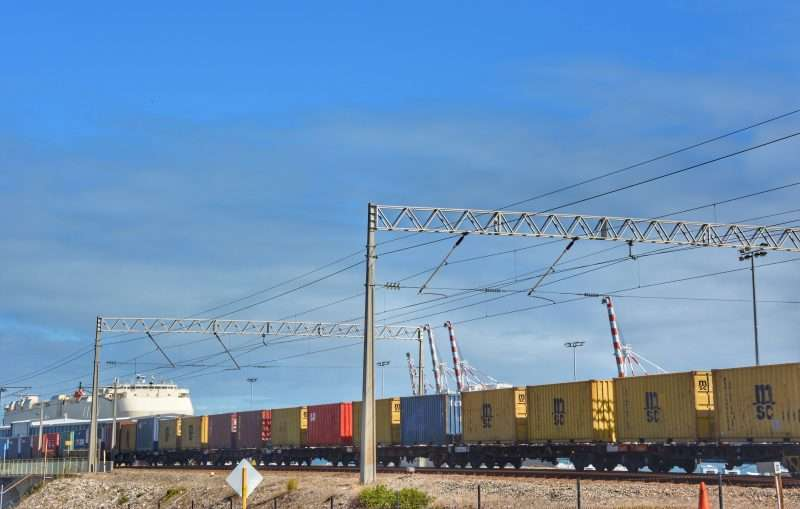Row of colorful shipping containers on a long freight train along the tracks under a clear blue sky with overhead power lines.